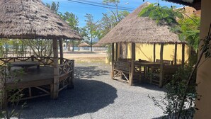 A covered outdoor seating area with thatched-roof structures, gravel ground, and surrounding greenery.