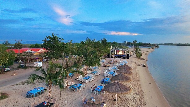 A beachside restaurant with outdoor seating, palm trees, and a view of the water.