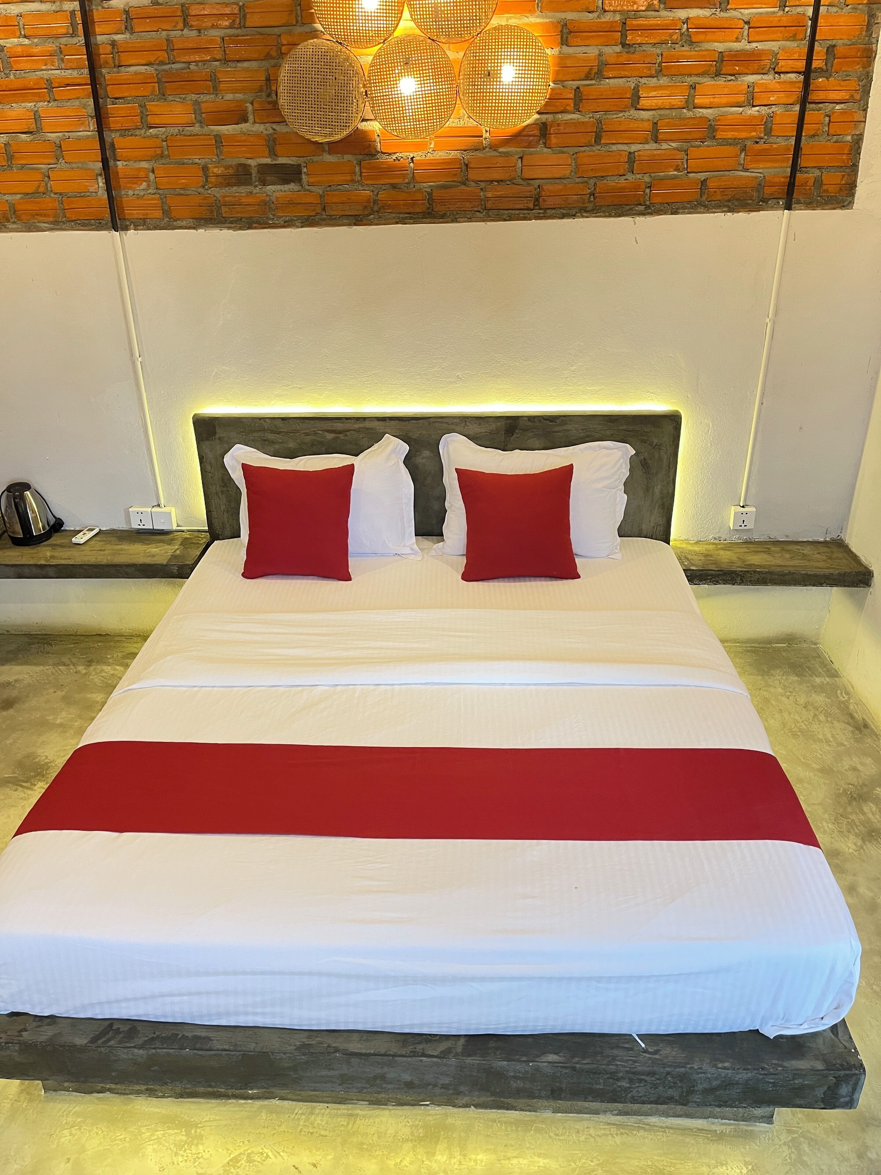 A bed with white and red bedding, a wooden headboard, and a brick wall with a hanging light fixture.