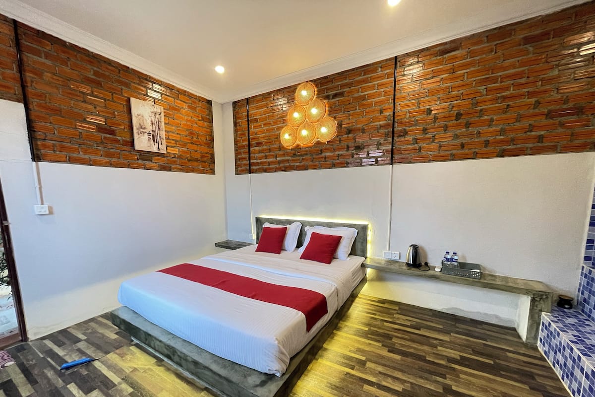 Une chambre moderne avec un mur en briques, un plancher en bois, un lit avec une literie blanche et rouge, une table de chevet avec une bouilloire et un luminaire fixé au mur.