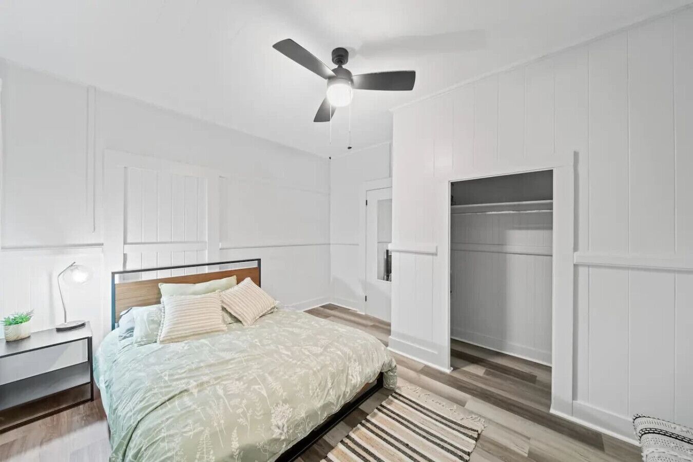 A bedroom with a bed, a ceiling fan, a nightstand, a lamp, a closet, and a wooden floor.