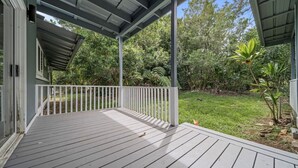 A covered wooden deck with a white railing, surrounded by a lush garden and trees.