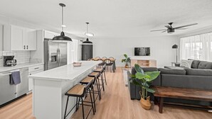 A modern kitchen with white cabinets, a large island with bar stools, a dining area with chairs, a grey sofa, and a TV on the wall.