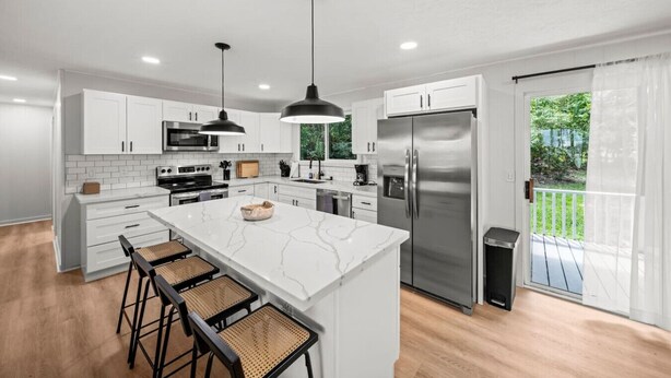 A modern kitchen with white cabinetry, a large island with bar stools, stainless steel appliances, and a view of a green garden through a sliding glass door.