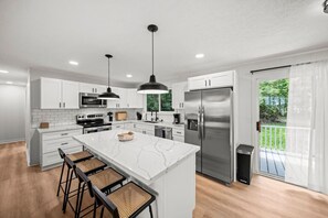 Une cuisine moderne avec des meubles blancs, un grand îlot central doté de tabourets, des appareils en acier inoxydable et une vue sur un jardin verdoyant grâce à une porte-fenêtre coulissante.