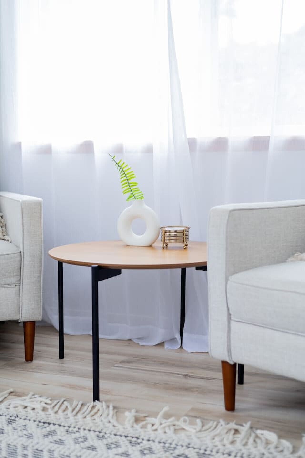 A room with a round wooden table, two armchairs, a white curtain, and a patterned rug.