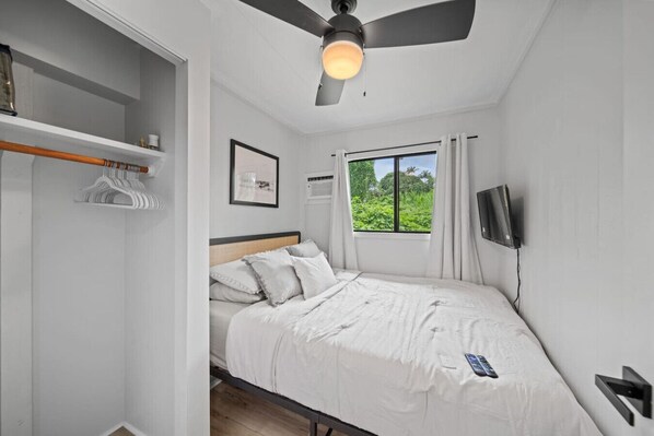 A bedroom with a bed, a ceiling fan, a window with a view of greenery, a TV mounted on the wall, and a wardrobe with hanging clothes.
