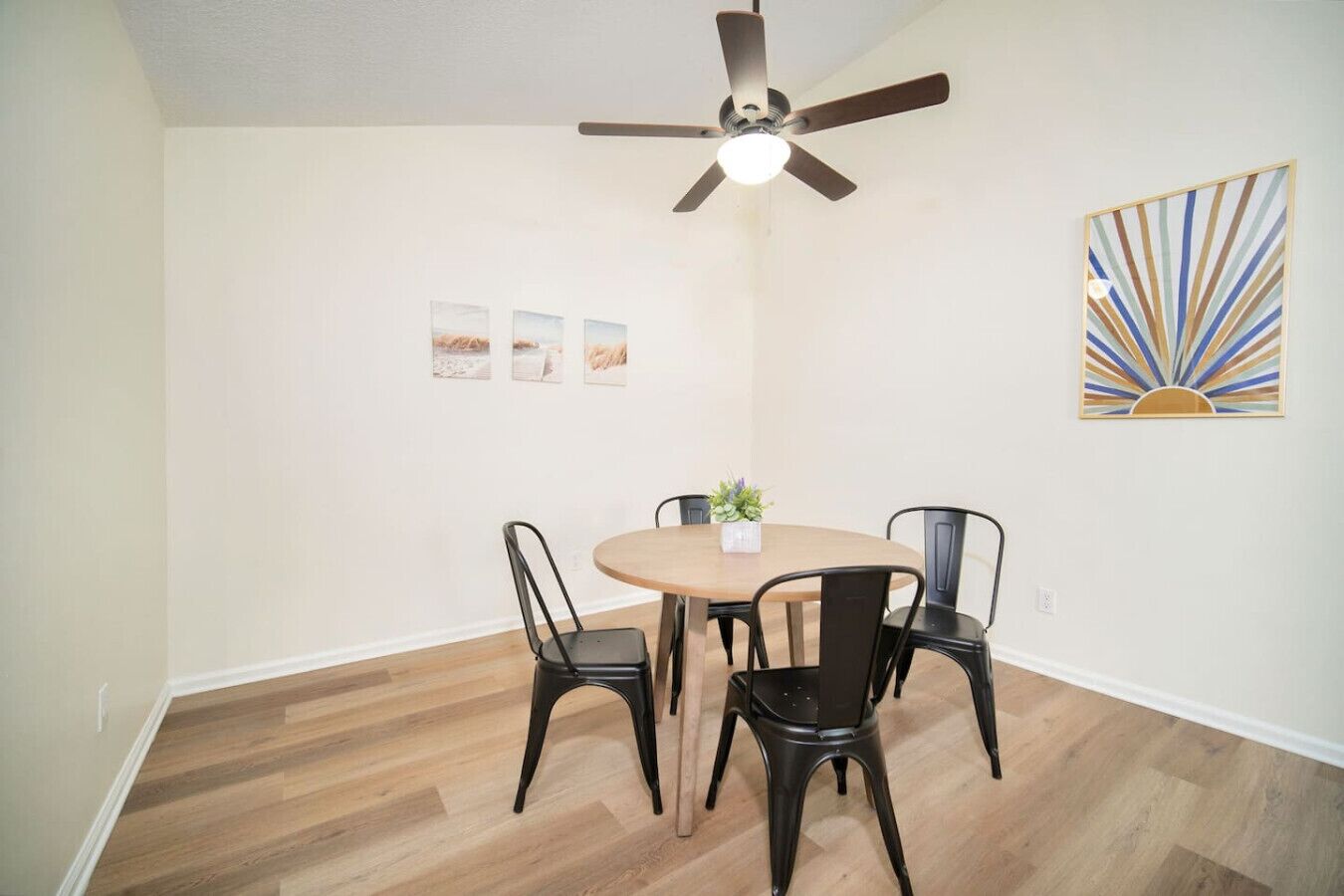 Un comedor con una mesa redonda, cuatro sillas, un ventilador de techo y una obra de arte colgada en la pared.
