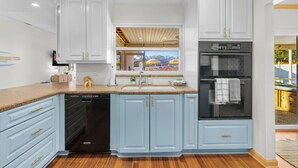 A kitchen with light blue cabinets, a dishwasher, a refrigerator, and a window with a view of outdoor umbrellas. - Private Resort | Pool, Spa, Tiki Bar + Games (Seminole)