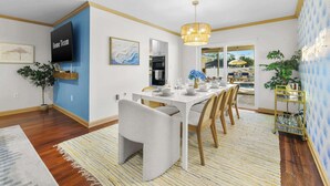 A dining area with a white table set for six, surrounded by chairs. There is a television mounted on the wall, a potted plant, and a wall with a blue pattern. - Private Resort | Pool, Spa, Tiki Bar + Games (Seminole)