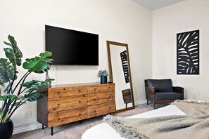 A modern living room with a wooden dresser, a flat-screen TV, a large mirror, a black leather chair, and a potted plant.