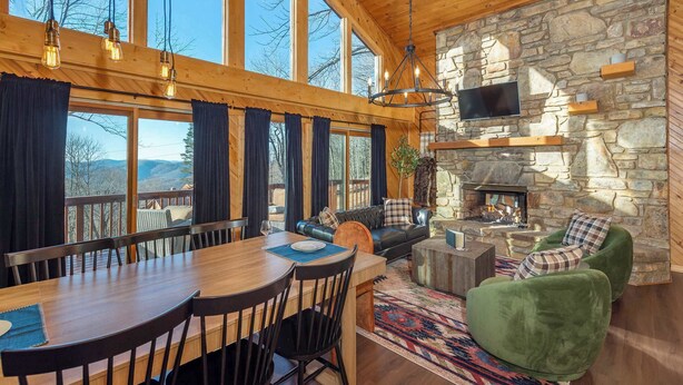 A spacious living area with a stone fireplace, wooden ceiling, and large windows overlooking a scenic landscape. - 6 Br Cabin~Hot Tub~Fire Pit~Min to Hatley Pointe (Mars Hill)