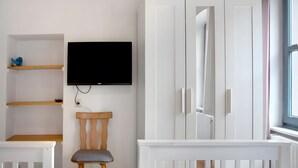 A room with a white crib, a wooden chair, a TV mounted on the wall, and a wardrobe with a mirror. - Holiday Apartment 'Alte Stadtmauer' with Shared Garden and Wi-Fi (Bamberg)