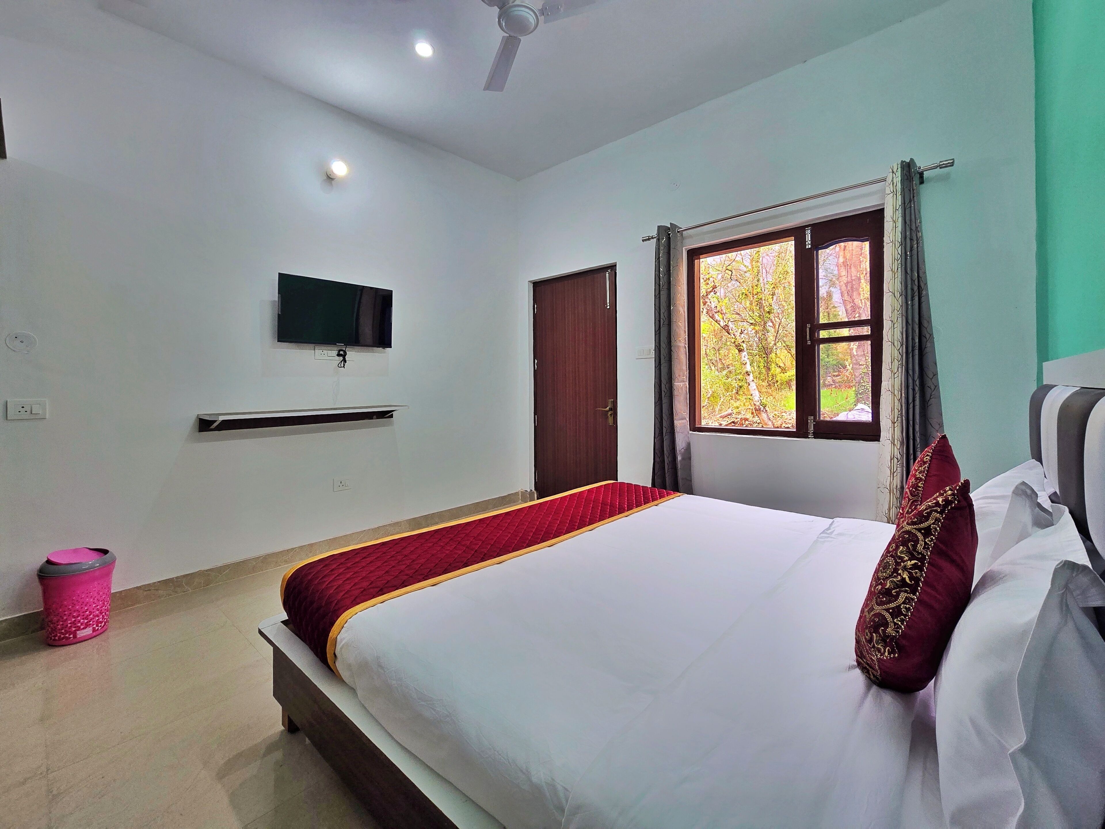 A bedroom with a bed, a TV mounted on the wall, a window with curtains, and a ceiling fan.