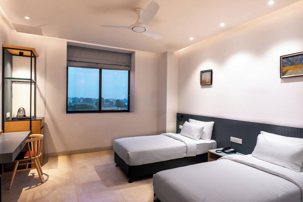 Ein modernes Hotelzimmer mit zwei Betten, einem Schreibtisch, einem Stuhl und einem großen Fenster mit Blick auf die Stadt.