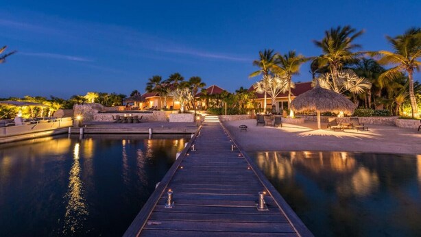 Een resort met een houten steiger naar een zwembad, omringd door palmbomen en constructies met rieten daken.