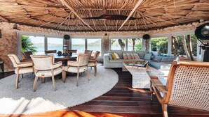 Een ruime kamer met rieten dak, een ronde eettafel met stoelen, een bank en een groot raam met uitzicht op zee.