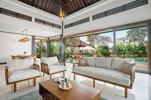 A spacious living area with a large sofa, two armchairs, a wooden coffee table, and a dining table with chairs. Large windows provide a view of the outdoor pool and greenery.
