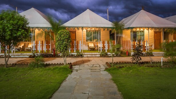 A row of three white-roofed buildings with a stone pathway leading to them, surrounded by greenery. - The Rawla Tent Resort & Spa (Garudeshwar)