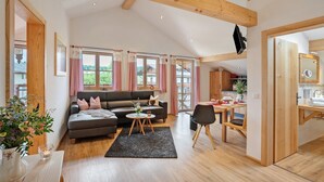Ein großzügiger Wohnbereich mit Sofa, Couchtisch und Esstisch, Holzbalken an der Decke, großen Fenstern mit Vorhängen sowie angrenzender Küche, die durch eine offene Tür sichtbar ist.