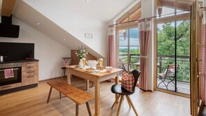 A dining area with a wooden table, bench, and chairs, a kitchen with an oven, and a balcony with a view. - Apartment 'Neubau - Freundliche' with Mountain View, Shared Garden and Wi-Fi (Höslwang)