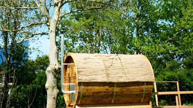 Une structure en forme de tonneau, faite de bois, dotée d’une porte en métal et entourée de verdure et d’arbres.