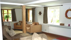 Un salon comprenant un canapé, des fauteuils, une colonne en bois, un radiateur, une fenêtre avec des rideaux, une plante en pot et un tapis coloré.