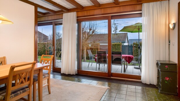 Een eetruimte met een houten tafel en stoelen, een glazen schuifdeur naar een terras, en een kastje met een bloemistisch ontwerp.