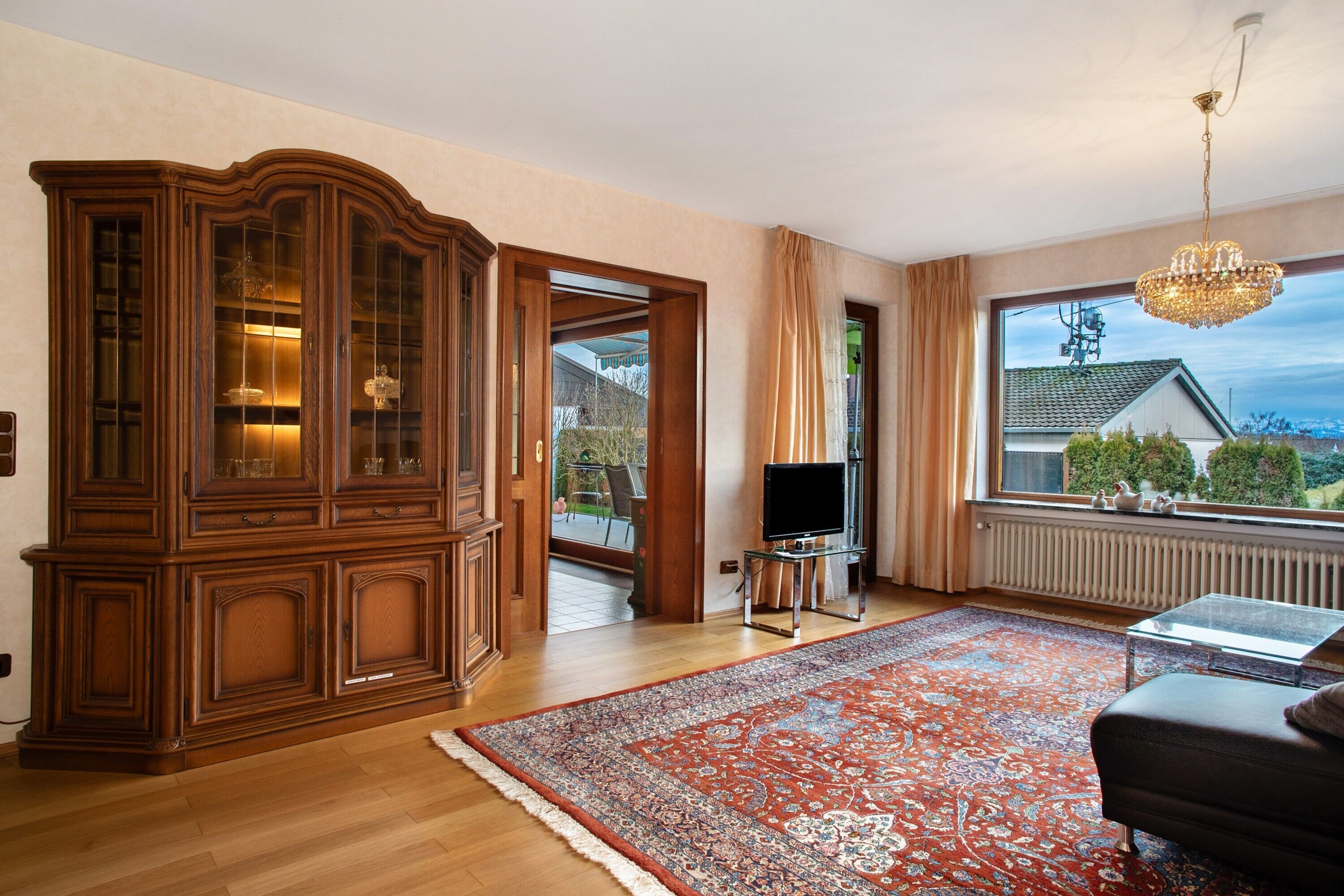 Ein Zimmer mit einem Holzkoffer, einem Fernseher auf einem Tisch, einem gemusterten Teppich und einem Fenster mit Blick auf ein Haus.