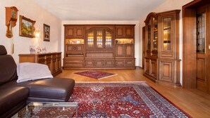 Een woonkamer met een bruine leren bank, een glazen salontafel, een rood tapijt met patroon, houten kasten en een kroonluchter.