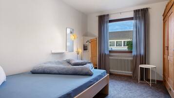 Ein Schlafzimmer mit Bett, Heizkörper, Fenster mit Vorhängen, einem kleinen Tisch und einem Kleiderschrank.