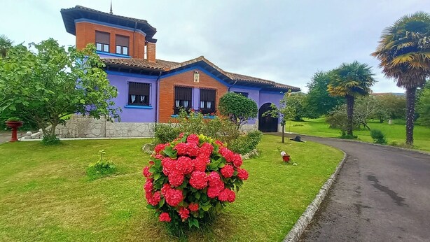 A two-story house with a brick facade and a blue trim, surrounded by a well-maintained lawn and flowering plants.