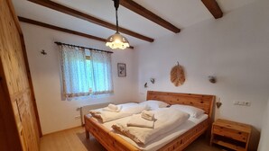 Uma cama de madeira com roupa de cama branca, uma pequena mesa de cabeceira, um tapete fofo e uma janela com cortinas estampadas.