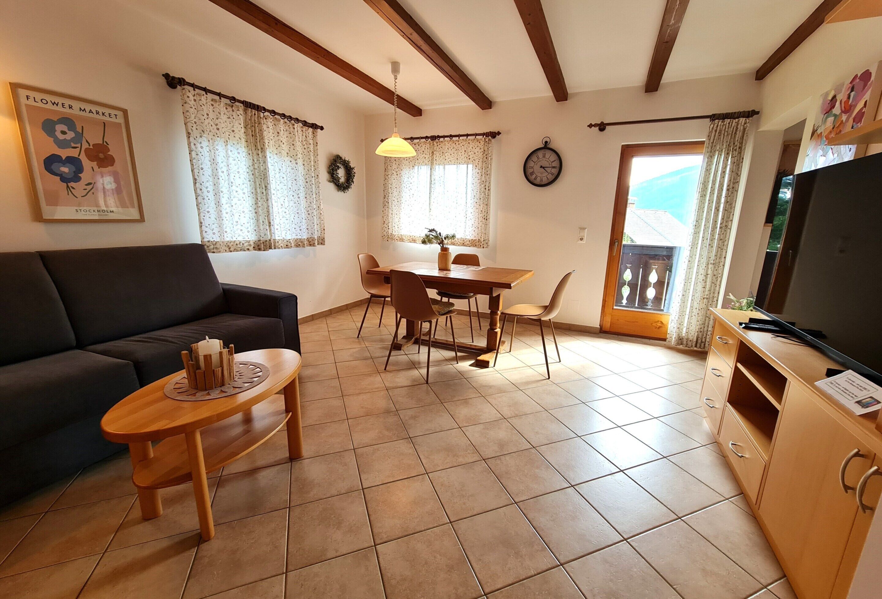 Una sala de estar con un sofá, una mesa de centro de madera, una mesa de comedor con sillas, un televisor y un reloj en la pared.