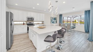 A modern kitchen with white cabinets, a central island with bar stools, stainless steel appliances, and pendant lighting. - LAKE VILLA 6bd/all un-suite, pool*spa*grill (Kissimmee)