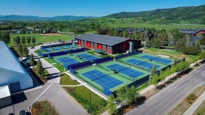 Tenniscomplex met meerdere banen, een groot gebouw met rode en zwarte gevel en een bergachtig landschap op de achtergrond.