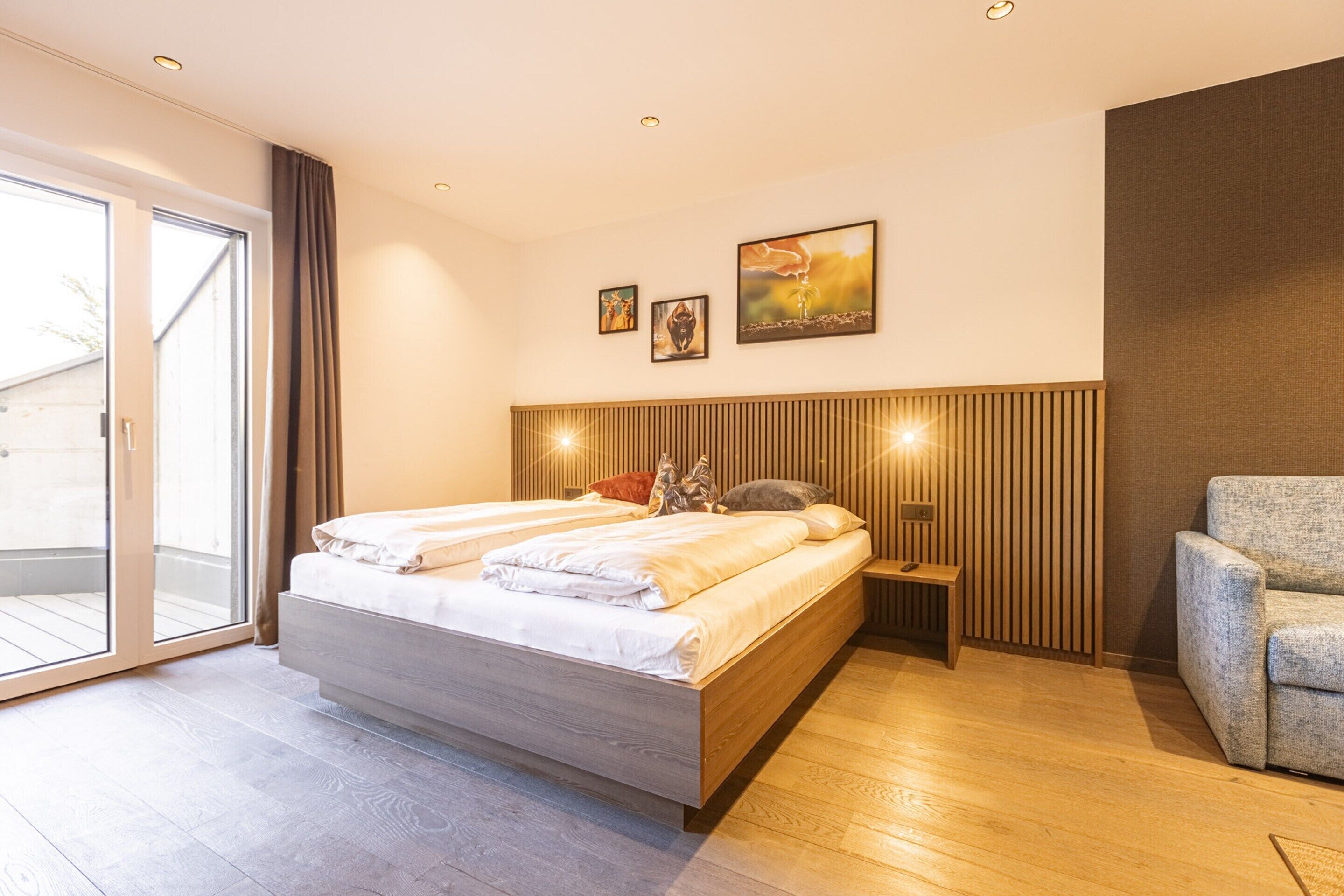 A modern bedroom with a large bed, a wooden headboard, a small bedside table, and a grey armchair.