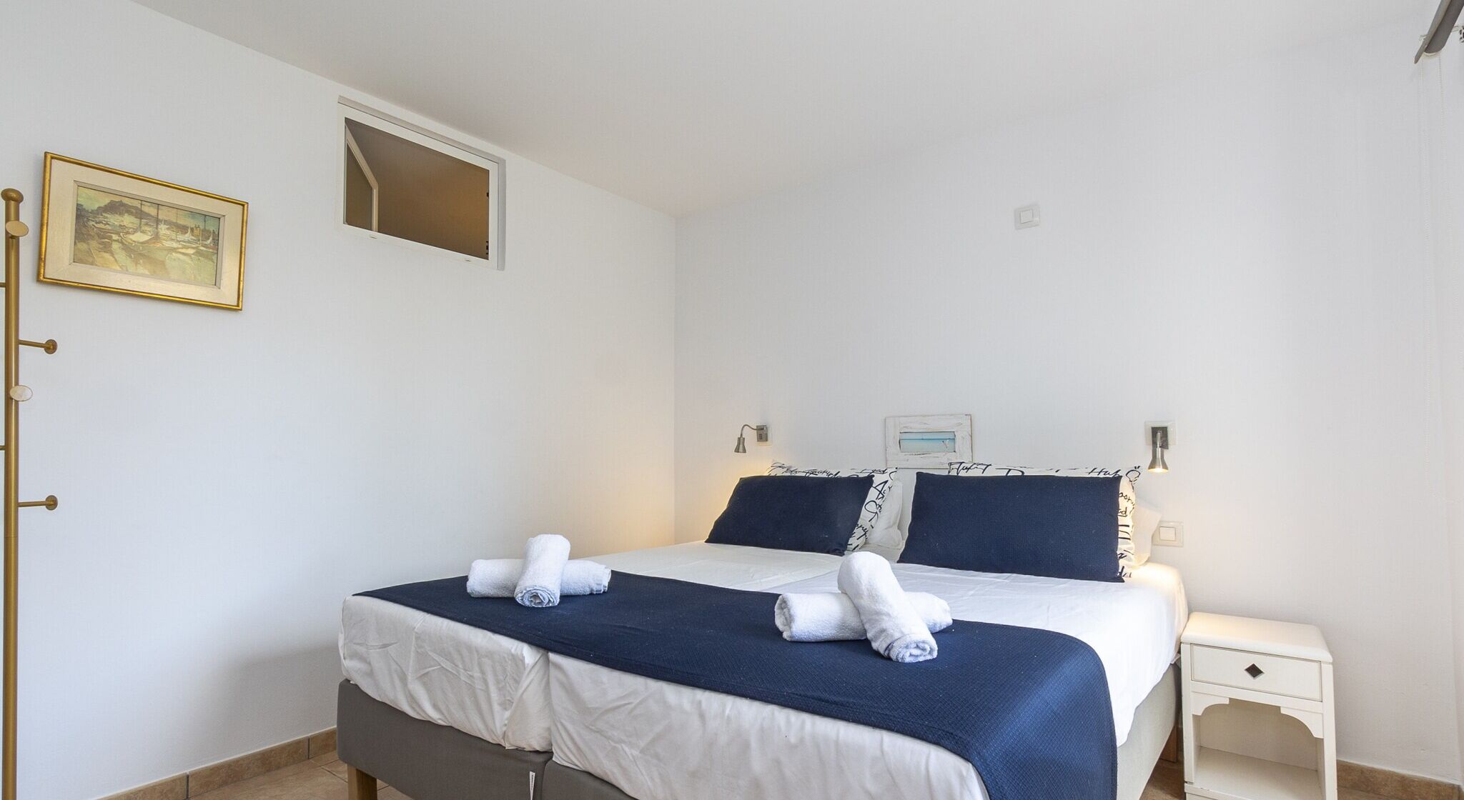 A neatly made bed with white and navy bedding, a small bedside table with a lamp, and a framed picture on the wall.