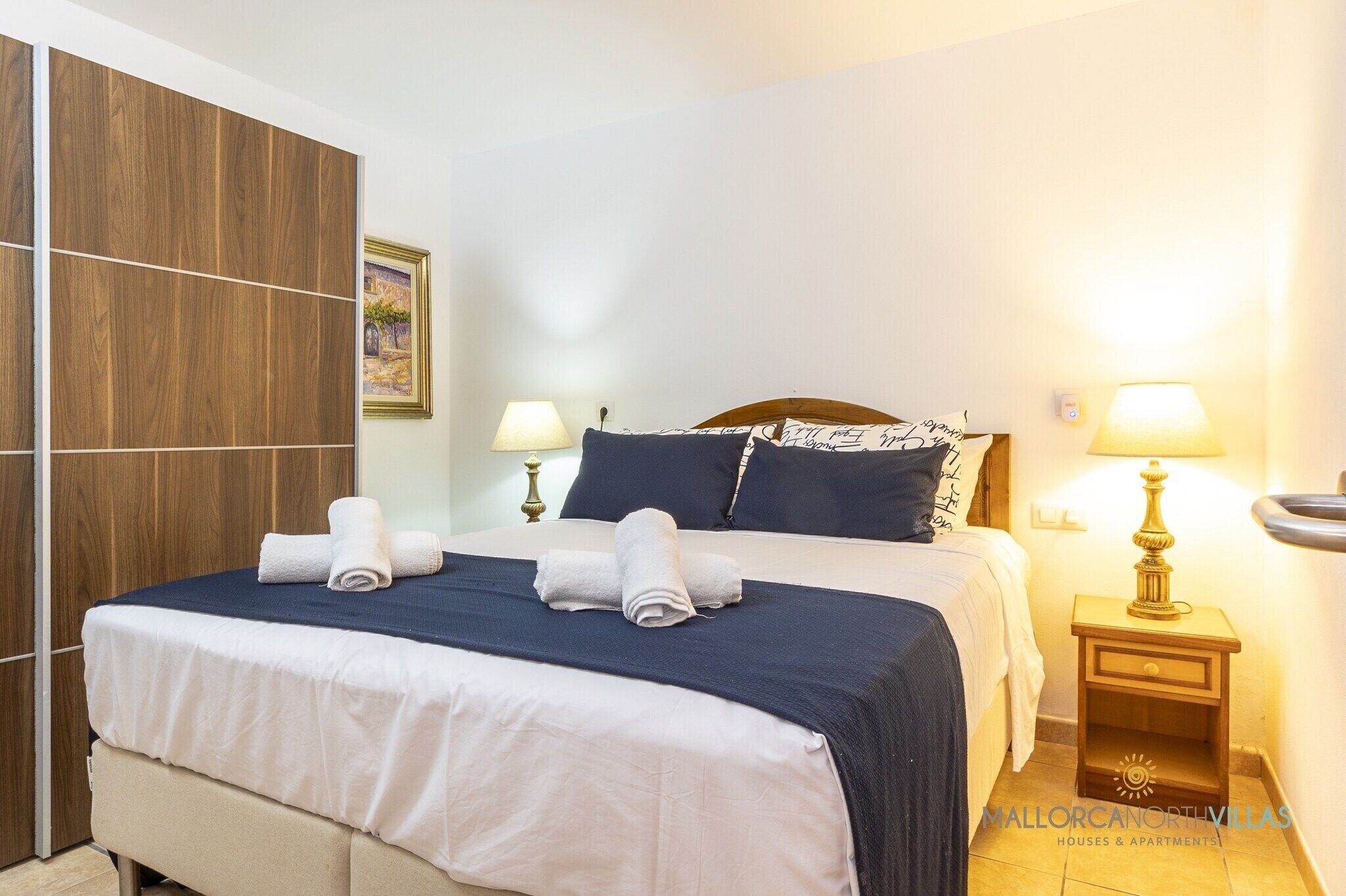 Une chambre d’hôtel avec un lit, deux lampes de chevet, une armoire en bois et un tableau encadré au mur.