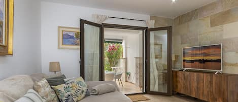 A living room with a sofa, a TV on a wooden stand, and a glass door leading to an outdoor area with a table and chairs.