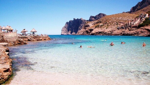 A beach with clear water, rocky terrain, and a few umbrellas. - House in Pollensa near Cala Barqués Beach (Pollensa)
