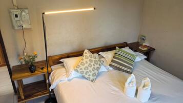 Ein ordentlich bezogenes Bett mit weißen Leinentüchern, einem hölzernen Kopfteil und dekorativen Kissen. Ein Nachttisch mit Blumen in einer Vase und einer Lampe sind ebenfalls vorhanden.