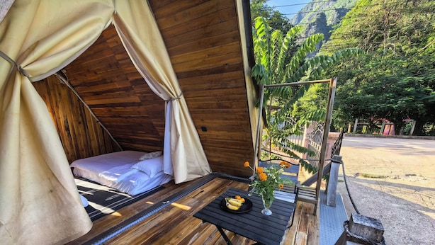 A wooden tent with a bed, a table with a vase of flowers, and a view of a road and greenery. - Mango Villa Nong Khiaw (Nong Khiaw)