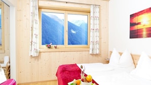 A hotel room with a wooden interior, a bed with white bedding, a window with a mountain view, and a breakfast tray with fruits and a cup.