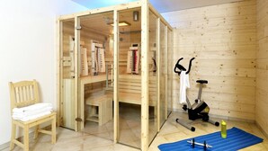 A wooden sauna room with a glass door, a wooden bench, a chair with towels, an exercise bike, and a blue exercise mat.
