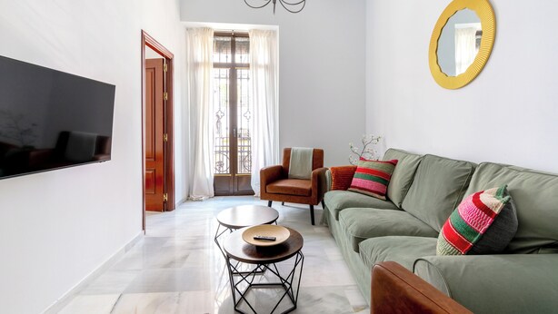 Una sala de estar con televisión de pantalla plana, un sofá, una butaca, una mesa redonda y un espejo en la pared.