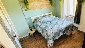 A bedroom with a wooden headboard, a floral bedspread, a small wooden stool, and a hanging light fixture. - On grassland (Malmerspach)