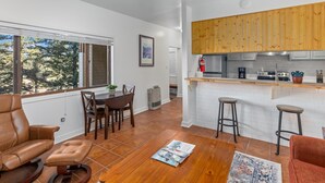 Una cucina moderna con mobili in legno, un piano di lavoro bianco e un fornello integrato. È presente un'area pranzo con tavolo e sedie, e un'ampia finestra che offre vista sugli alberi.