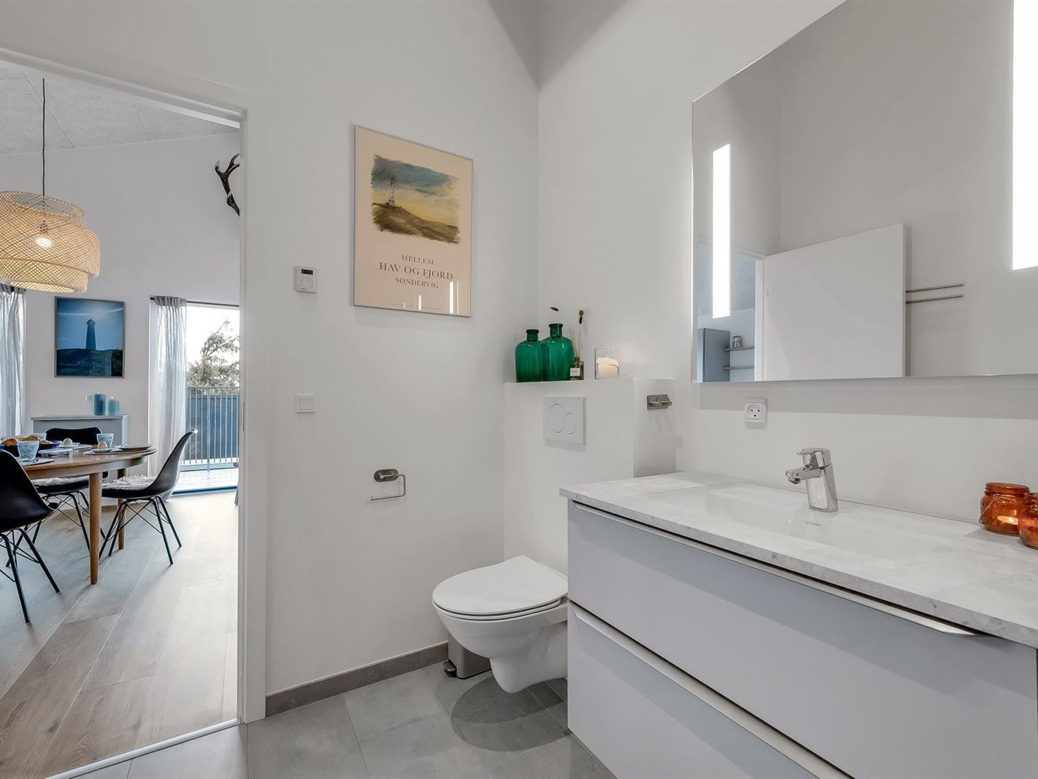 A modern bathroom with a toilet, sink, and mirror. There is a framed picture on the wall and a bottle of green liquid on the counter.