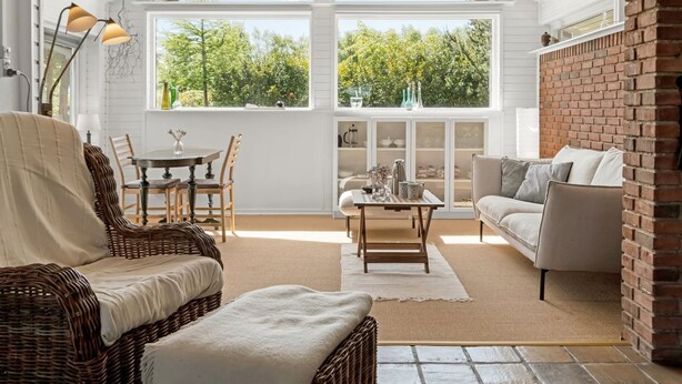 Ein geräumiges Wohnzimmer mit hoher Decke, großen Fenstern, einer Ziegelwand und einer gemütlichen Sitzecke mit Sofa und Korbsessel.